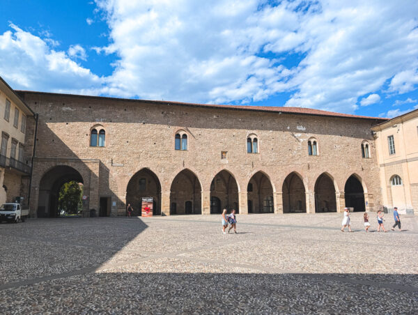 Piazza della Cittadella in Bergamo shows a long stone arcade with arched openings under a bright blue sky as people cross the cobbled square.