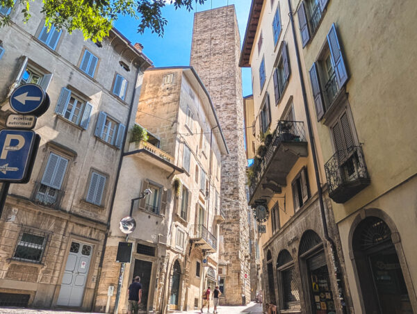 Pedestrians walk along Via Colleoni in Bergamo, framed by tall historic buildings and a church tower rising in the distance.