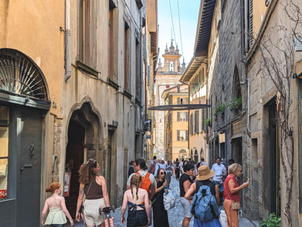 Tourists walk along Via Colleoni in Bergamo, flanked by old stone and stucco buildings beneath a distant church tower on a sunny day.